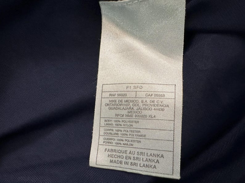 May include: A close-up of a white label with black text, attached to a navy blue fabric. The label includes the text "F1 SFD", "RN 58023", and "CA# 05553". It also lists the manufacturer's address and material composition.