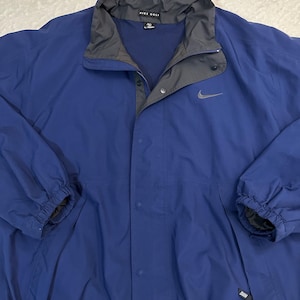 May include: Blue Nike Golf jacket with a gray collar and a gray Nike swoosh logo. The jacket has a button-up front, two side pockets, and elastic cuffs. The jacket is made of a lightweight material.