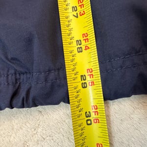 May include: Close-up of navy blue fabric with a yellow measuring tape. The tape shows measurements from 68.58 cm to 76.2 cm. The fabric appears to be part of a garment, possibly a jacket or coat.