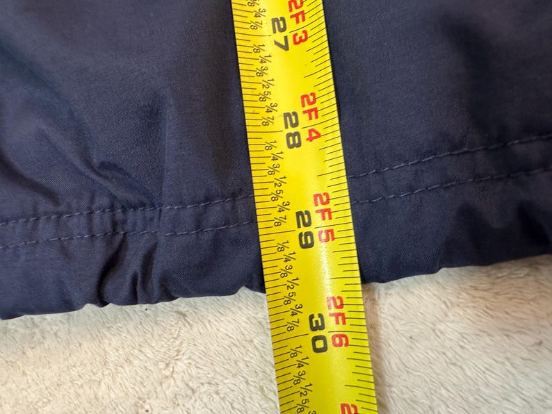 May include: Close-up of navy blue fabric with a yellow measuring tape. The tape shows measurements from 68.58 cm to 76.2 cm. The fabric appears to be part of a garment, possibly a jacket or coat.