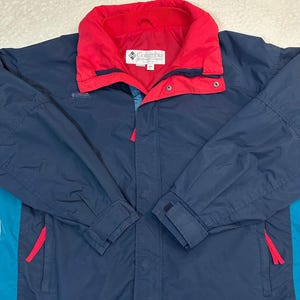 May include: A navy blue Columbia jacket with a red collar and accents. The jacket features a teal side panel and red zipper pulls. The Columbia logo is visible on the chest. The jacket has a zip closure and adjustable cuffs.
