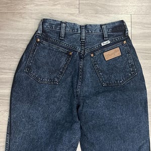 May include: Dark wash denim jeans with a high waist and two back pockets. The Wrangler brand label is visible on the back pocket. The jeans have a classic five-pocket design and a button and zipper closure.