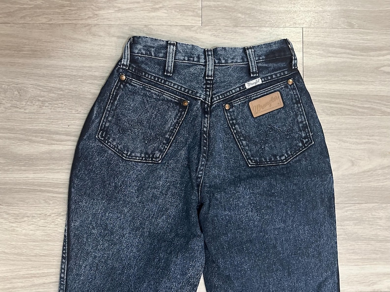 May include: Dark wash denim jeans with a high waist and two back pockets. The Wrangler brand label is visible on the back pocket. The jeans have a classic five-pocket design and a button and zipper closure.