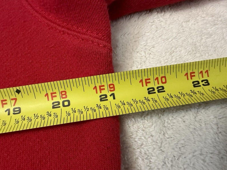 Puede incluir: Una cinta m&eacute;trica amarilla con marcas negras se coloca sobre una superficie de tela roja. La cinta mide hasta 58,4 cm.