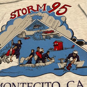 May include: A light gray t-shirt featuring a graphic of a storm scene with the text "STORM 95" in red. The illustration depicts people in boats and buildings in a flood. The text "MONTECITO CA" is at the bottom.