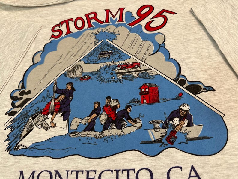 May include: A light gray t-shirt featuring a graphic of a storm scene with the text "STORM 95" in red. The illustration depicts people in boats and buildings in a flood. The text "MONTECITO CA" is at the bottom.