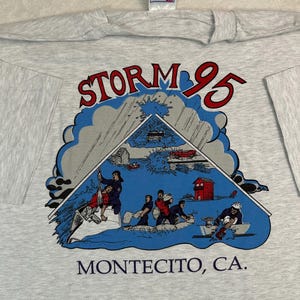 May include: A light gray t-shirt featuring a graphic of a storm scene with the text "STORM 95" in red and blue. The graphic includes illustrations of people, buildings, and vehicles. The text "MONTECITO, CA." is printed below the graphic.