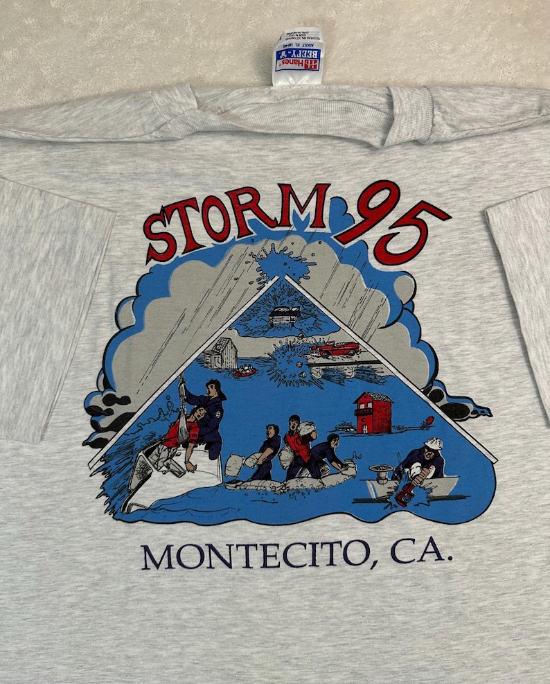May include: A light gray t-shirt featuring a graphic of a storm scene with the text "STORM 95" in red and blue. The graphic includes illustrations of people, buildings, and vehicles. The text "MONTECITO, CA." is printed below the graphic.