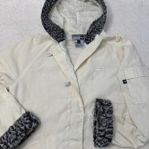 May include: A white hooded jacket with a grey fleece lining. The jacket has a zip closure and button accents. The jacket has a logo on the left chest that says "ROXY".