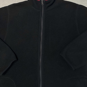 Puede incluir: Chaqueta polar negra con cremallera completa y forro interior rojo visible en el cuello. La chaqueta tiene cuello alto y dos bolsillos delanteros. Las mangas son largas y el material parece suave.