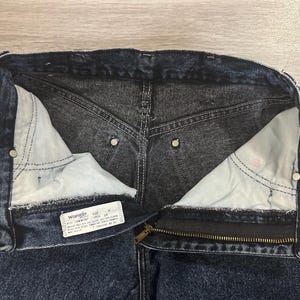 May include: Close-up of dark wash denim jeans with a partially unzipped zipper. The interior pockets are visible, along with the Wrangler label. The jeans have a classic five-pocket design and metal rivets.