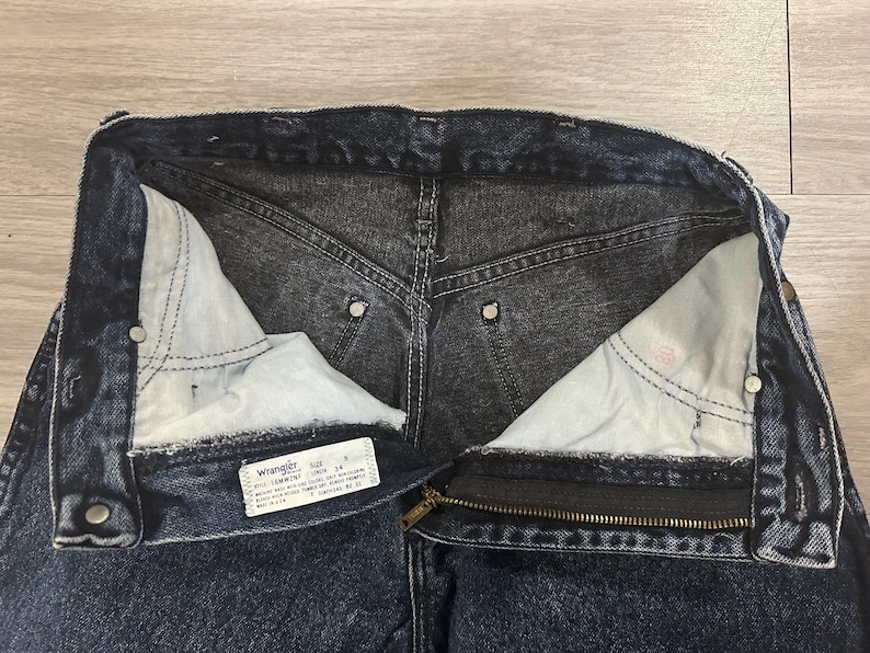 May include: Close-up of dark wash denim jeans with a partially unzipped zipper. The interior pockets are visible, along with the Wrangler label. The jeans have a classic five-pocket design and metal rivets.