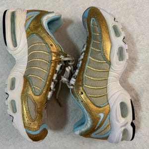 Pu&ograve; includere: Paio di scarpe sportive dorate, beige e azzurro. Le scarpe hanno una suola bianca con un accento nero e un interno azzurro. Le tomaie sono dorate con dettagli beige e azzurri. I lacci sono bianchi.