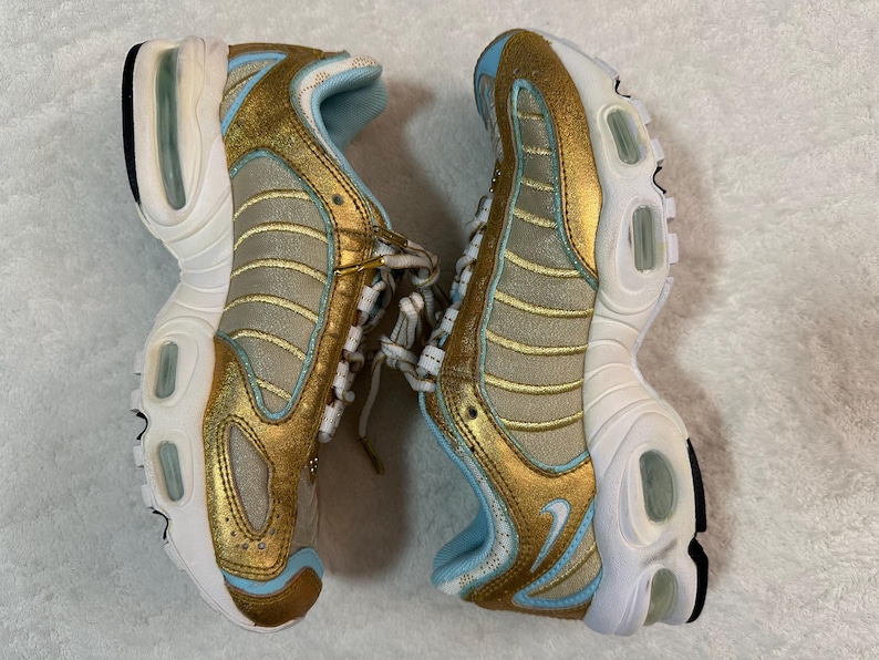 Pu&ograve; includere: Paio di scarpe sportive dorate, beige e azzurro. Le scarpe hanno una suola bianca con un accento nero e un interno azzurro. Le tomaie sono dorate con dettagli beige e azzurri. I lacci sono bianchi.