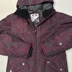 Può includere: Una giacca da sci a fantasia bordeaux e nera con chiusura a zip e cappuccio. La giacca ha una patch con il logo sul petto sinistro che dice "Oakley" e "Modular 3L".