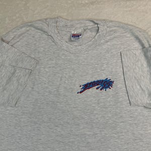 May include: A light gray t-shirt with short sleeves. The shirt features a small graphic on the left chest that reads "STORM 92" in blue and red. The shirt is made of a soft material and has a crew neck.