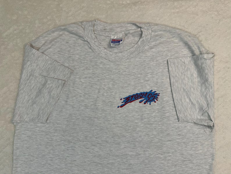 May include: A light gray t-shirt with short sleeves. The shirt features a small graphic on the left chest that reads "STORM 92" in blue and red. The shirt is made of a soft material and has a crew neck.