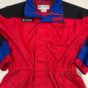 May include: A red, blue, and black Columbia Sportswear Company jacket. The jacket features a blue collar and sleeve accents, with the brand logo visible. The word "TECTANIUM" is printed on the chest pocket. It has a drawstring waist and a full-length zip.
