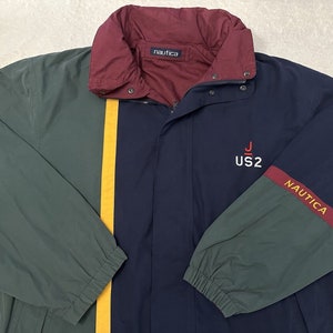 Op de afbeelding: Een veelkleurige Nautica-jas in groen, marineblauw en bordeauxrood. De jas heeft een gele streep en het Nautica-logo op de mouw. Op de borst staat de tekst "US2" en de letter "J".