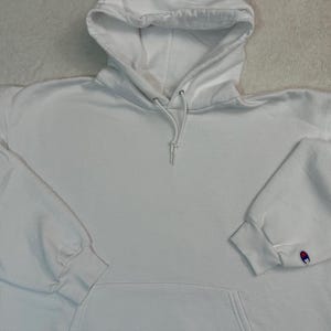 Puede incluir: Una sudadera con capucha Champion blanca con capucha con cordón y bolsillo de canguro. Las mangas largas tienen puños acanalados y el logotipo de Champion es visible en el puño izquierdo. La sudadera con capucha está hecha de un material suave y cómodo.