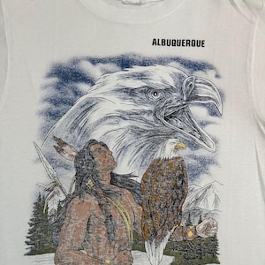 Puede incluir: Una camiseta blanca con un estampado gráfico de una persona con pelo largo y oscuro, una lanza y un águila calva posada en el brazo. Arriba hay una ilustración grande y detallada de una cabeza de águila y el texto ALBUQUERQUE.