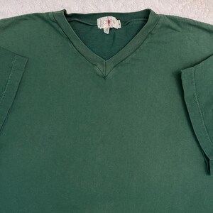 May include: A solid dark green short-sleeved t-shirt with a V-neck collar. The shirt features a ribbed neckline and a white fabric tag at the centre of the collar with the text J.CREW printed in black letters. The fabric has a matte finish.