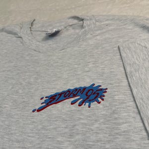 May include: A heather gray long-sleeve t-shirt with the text "STORM 95" in red and blue. The graphic has a splatter design. The shirt is made of a soft material.