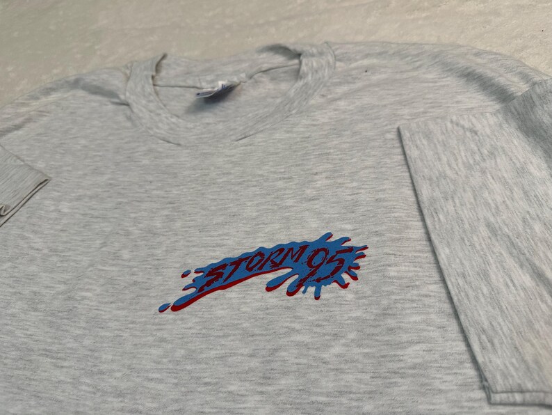 May include: A heather gray long-sleeve t-shirt with the text "STORM 95" in red and blue. The graphic has a splatter design. The shirt is made of a soft material.