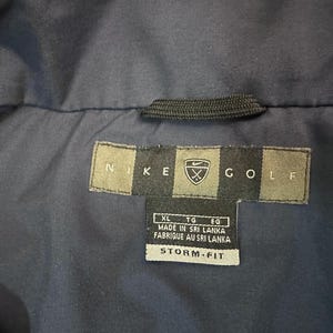 May include: A close-up of a navy blue jacket featuring a Nike Golf label. The label is rectangular with the Nike logo and the word "GOLF". The label also includes text indicating the jacket is made in Sri Lanka and is Storm-Fit.