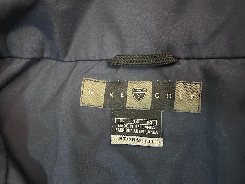May include: A close-up of a navy blue jacket featuring a Nike Golf label. The label is rectangular with the Nike logo and the word "GOLF". The label also includes text indicating the jacket is made in Sri Lanka and is Storm-Fit.