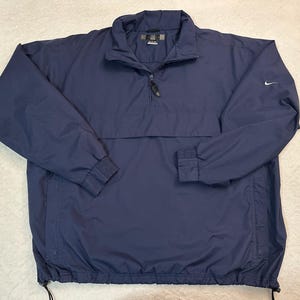 May include: A navy blue Nike pullover jacket with a quarter-zip, a stand-up collar, and elasticated cuffs. The jacket features a small white Nike swoosh logo on the left sleeve. The hem has an adjustable drawstring. This lightweight jacket is suitable for outdoor activities.