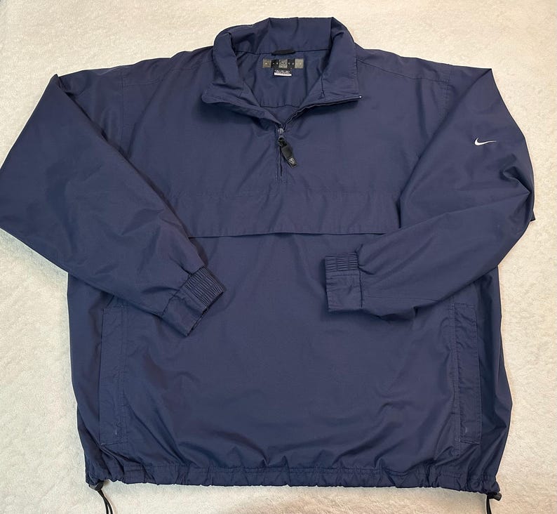 May include: A navy blue Nike pullover jacket with a quarter-zip, a stand-up collar, and elasticated cuffs. The jacket features a small white Nike swoosh logo on the left sleeve. The hem has an adjustable drawstring. This lightweight jacket is suitable for outdoor activities.