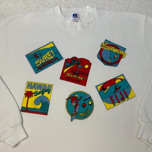 May include: White long-sleeve sweatshirt with colorful patches featuring surfing locations and the words "Surf! Australia", "Malibu Surfing", "Hawaii Surfing", "Fiji", and "Maui".