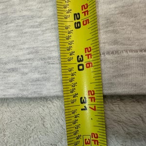 May include: A close-up shot of a gray fabric with a yellow and black measuring tape. The tape shows measurements in inches, with the numbers 29, 30, and 31 clearly visible. The fabric has a textured appearance.