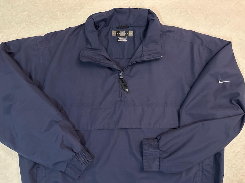 May include: Navy blue pullover jacket with a quarter-zip and a small white Nike logo on the sleeve. The jacket has a stand-up collar and elastic cuffs. The jacket is made of a lightweight, water-resistant material.