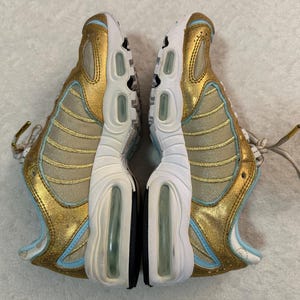 Pu&ograve; includere: Paio di scarpe sportive oro e azzurro chiaro. Le scarpe da ginnastica hanno una suola bianca con cuscinetti d'aria e un accento nero. Le tomaie sono dorate con accenti azzurri e lacci bianchi. Vista dall'alto.