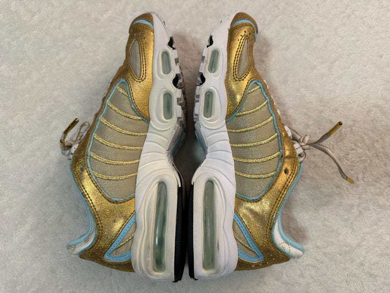 Pu&ograve; includere: Paio di scarpe sportive oro e azzurro chiaro. Le scarpe da ginnastica hanno una suola bianca con cuscinetti d'aria e un accento nero. Le tomaie sono dorate con accenti azzurri e lacci bianchi. Vista dall'alto.