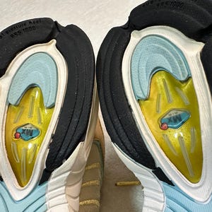 Pu&ograve; includere: Primo piano delle suole di due scarpe sportive. Le suole presentano un tacco in gomma nero, un interno azzurro e un centro giallo con la scritta "MX AIR". Le scarpe hanno un esterno bianco e beige.