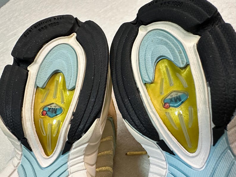 Pu&ograve; includere: Primo piano delle suole di due scarpe sportive. Le suole presentano un tacco in gomma nero, un interno azzurro e un centro giallo con la scritta "MX AIR". Le scarpe hanno un esterno bianco e beige.