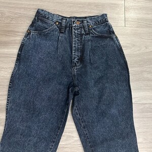 May include: High-waisted, acid-wash denim jeans. The jeans have a classic five-pocket design, a button and zipper closure, and brass-colored rivets. The denim has a faded, mottled appearance, and the jeans are in good condition.