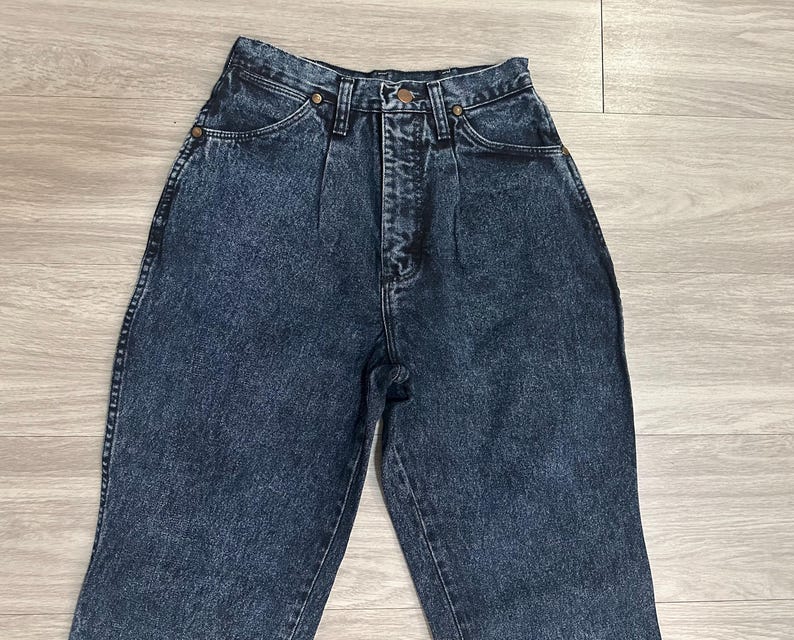 May include: High-waisted, acid-wash denim jeans. The jeans have a classic five-pocket design, a button and zipper closure, and brass-colored rivets. The denim has a faded, mottled appearance, and the jeans are in good condition.