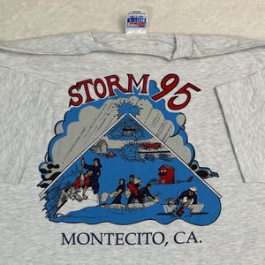 May include: A light gray t-shirt featuring a graphic of a storm scene with the text "STORM 95" in red and "MONTECITO, CA." in black. The graphic depicts people in a boat and a red building.