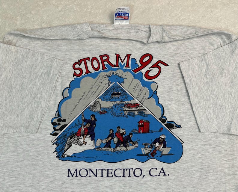 May include: A light gray t-shirt featuring a graphic of a storm scene with the text "STORM 95" in red and "MONTECITO, CA." in black. The graphic depicts people in a boat and a red building.