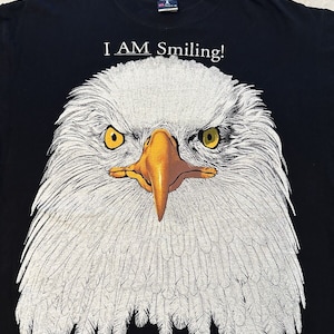 Könnte beinhalten: Schwarzes T-Shirt mit einem weißen Kopf eines Weißkopfseeadlers. Der Adler hat gelbe Augen und einen gelben Schnabel. Der Text "I AM Smiling!" befindet sich über dem Adler und "ALASKA RAPTOR CENTER Sitka, Alaska" unter dem Adler.