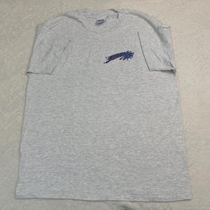 May include: A light gray t-shirt with short sleeves. The shirt features the word "STUDIO" in a stylized design on the upper left chest area. The text is in a combination of red and blue colors.