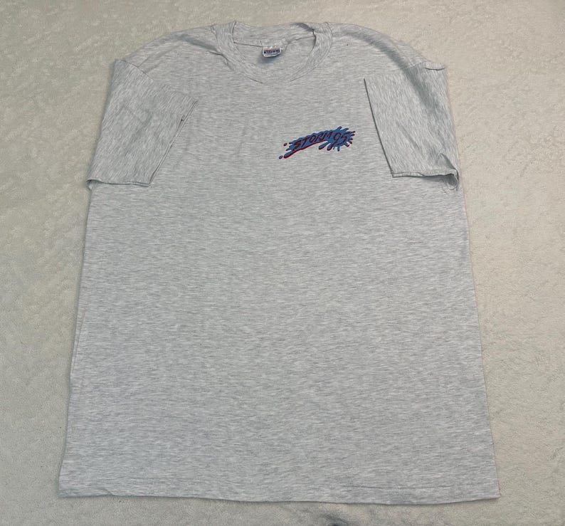 May include: A light gray t-shirt with short sleeves. The shirt features the word "STUDIO" in a stylized design on the upper left chest area. The text is in a combination of red and blue colors.