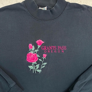 Puede incluir: Una sudadera negra de manga larga con cuello alto. La parte delantera presenta un diseño bordado de tres rosas de color rosa con tallos y hojas verdes, acompañado del texto bordado "GRANTS PASS OREGON" en letras de color rosa.