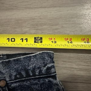 May include: Close-up of denim fabric with a yellow measuring tape. The tape shows measurements from 9 to 16 inches. The denim has a button and a rivet. The image is on a wooden surface.