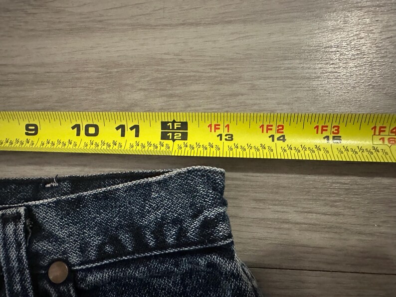 May include: Close-up of denim fabric with a yellow measuring tape. The tape shows measurements from 9 to 16 inches. The denim has a button and a rivet. The image is on a wooden surface.