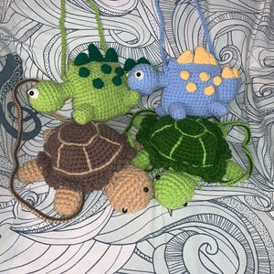 May include: Crocheted dinosaur and turtle-shaped crossbody bags in green, blue, brown, and tan. The bags have long straps and are designed with detailed features. The bags are handmade and suitable for children.